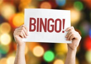 Zwei Hände halten ein Blatt Papier mit dem Wort Bingo vor unscharfen Lichtern